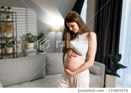Young woman cradles her pregnant belly with a gentle smile, standing in a softly lit living room with plants and a sofa 134805876