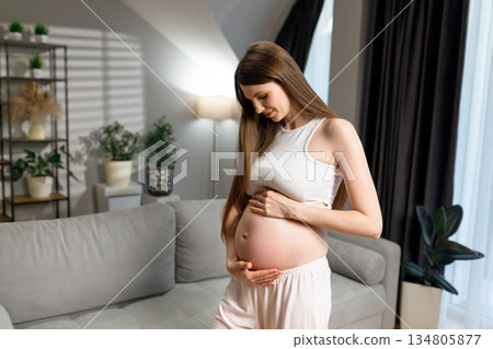 Young woman cradling her pregnant belly with a gentle smile, standing in a bright, modern living room with plants and a sofa Young woman cradling her pregnant belly with a gentle smile, standing in a bright, modern living room with plants and a sofa 134805877