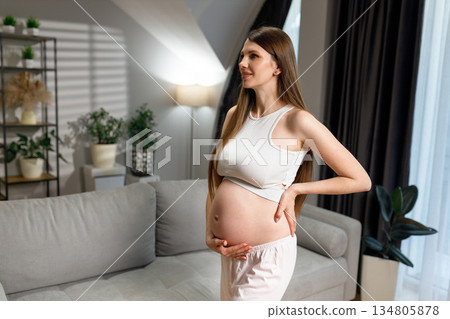Young expectant mother in casual wear stands indoors with hands on her belly, showing her pregnancy Young expectant mother in casual wear stands indoors with hands on her belly, showing her pregnancy 134805878