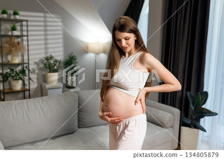 Young expectant mother stands in living room, cradling her belly with gentle smile, anticipating childbirth 134805879