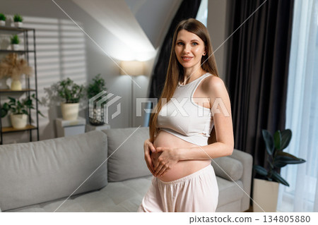 Pregnant woman in white top and pajama pants forms heart shape on belly with hands, smiling gently in cozy living room with plants and sofa 134805880