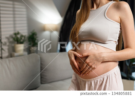 Pregnant woman cradles her belly forming a heart shape with her hands, indoors with soft lighting and home decor elements visible 134805881