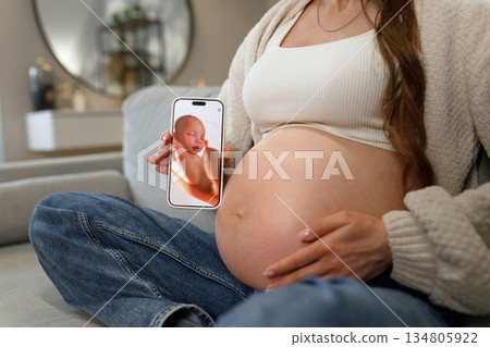 Pregnant woman holding smartphone displaying ultrasound image of her baby while sitting on couch, anticipation and joy evident Pregnant woman holding smartphone displaying ultrasound image of her baby while sitting on couch, anticipation and joy evident 134805922