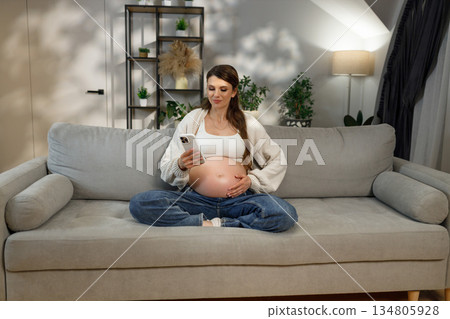 Expecting mother sits cross-legged on sofa, holding phone and touching belly, enjoying quiet moment at home 134805928