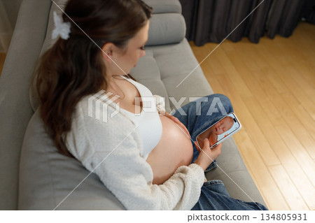 Pregnant woman sits on sofa holding smartphone displaying ultrasound image of her unborn baby, anticipating motherhood 134805931