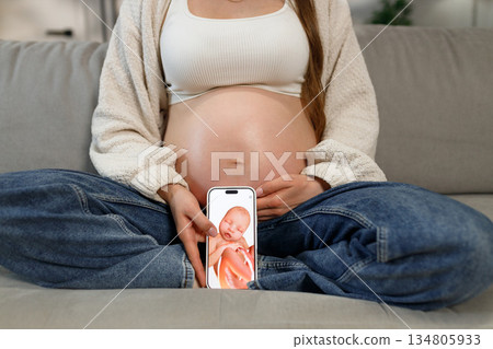 Pregnant woman sits on couch holding smartphone displaying 3D ultrasound of baby, showing anticipation and connection 134805933