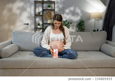 Pregnant woman sits on couch looking at phone displaying ultrasound image, showing anticipation and connection to unborn baby 134805935