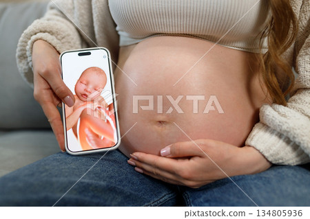 Pregnant woman holds smartphone displaying ultrasound image of fetus next to her belly, showcasing prenatal technology and anticipation 134805936