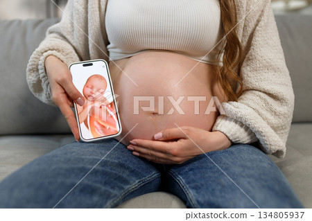 Pregnant woman holds smartphone displaying ultrasound image of fetus next to her belly, indoors on sofa 134805937