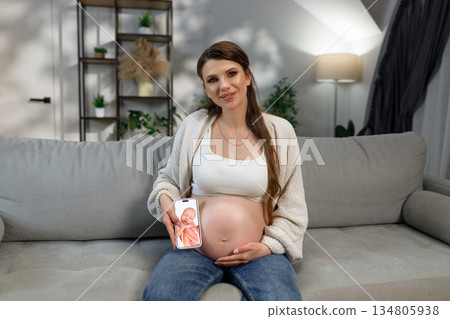 Expecting mother sits on sofa holding smartphone displaying ultrasound scan of baby, indoors with home decor visible 134805938