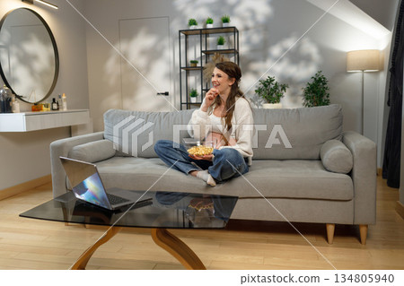 Young woman with long hair sits cross-legged on a gray sofa, holding a bowl of popcorn and eating it while watching something on a laptop Young woman with long hair sits cross-legged on a gray sofa, holding a bowl of popcorn and eating it while watching something on a laptop 134805940