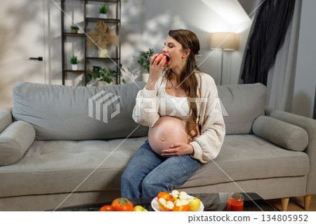 Expecting mother enjoys healthy snack of fresh apple while sitting on comfortable couch in living room, showcasing maternal well-being and nutrition Expecting mother enjoys healthy snack of fresh apple while sitting on comfortable couch in living room, showcasing maternal well-being and nutrition 134805952