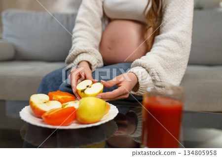 Pregnant woman holding apple slices with persimmons and juice nearby, focusing on healthy nutrition during pregnancy 134805958