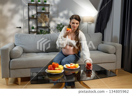 Pregnant woman sits on sofa, eating orange slice and holding glass of juice, with plates of fruit nearby Pregnant woman sits on sofa, eating orange slice and holding glass of juice, with plates of fruit nearby 134805962