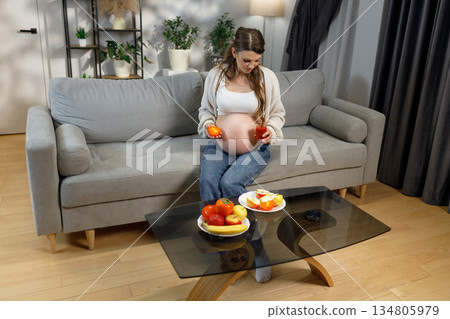 Pregnant woman sits on sofa holding orange and juice, with fruit on coffee table, healthy eating concept Pregnant woman sits on sofa holding orange and juice, with fruit on coffee table, healthy eating concept 134805979