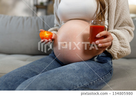 Pregnant woman in jeans and cozy sweater sits on sofa holding tomato juice and slice of tomato Pregnant woman in jeans and cozy sweater sits on sofa holding tomato juice and slice of tomato 134805980