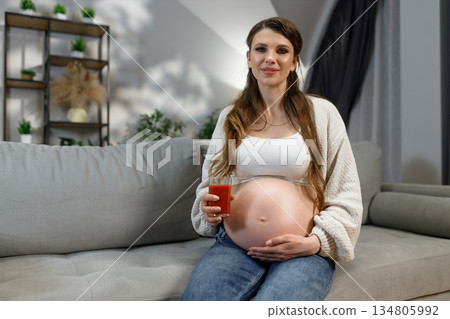 Pregnant woman sits on sofa holding glass of red juice, cradling belly, looking at camera with gentle smile 134805992