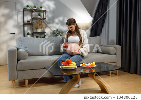 Pregnant woman sits on sofa holding glass of juice and touching belly, fruit on coffee table Pregnant woman sits on sofa holding glass of juice and touching belly, fruit on coffee table 134806125