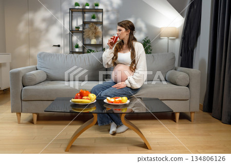 Pregnant woman sits on sofa drinking juice with fruit on table, expecting mother enjoying healthy beverage and snacks at home 134806126