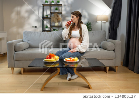 Pregnant woman sits on sofa drinking juice with fruit on table, expecting mother enjoying healthy refreshment at home 134806127