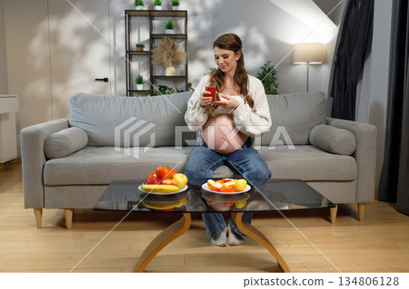 Pregnant woman sits on sofa holding glass of juice with fruit on table, expecting mother enjoying healthy refreshment at home 134806128
