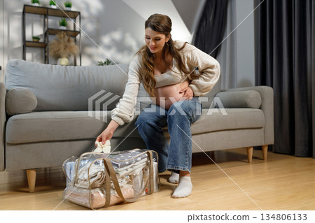 Expecting mother sits on sofa, packing baby clothes into transparent hospital bag, preparing for childbirth 134806133