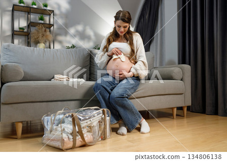 Expecting mother sits on sofa preparing baby clothes and hospital bag for childbirth Expecting mother sits on sofa preparing baby clothes and hospital bag for childbirth 134806138