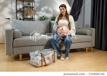 Expecting mother sits on sofa with packed hospital bag nearby, preparing for childbirth and new beginnings 134806140