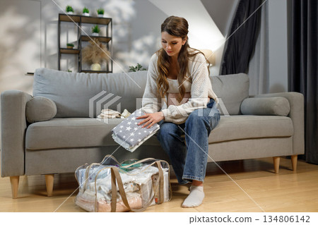 Expecting mother sits on sofa folding baby clothes preparing hospital bag for childbirth 134806142