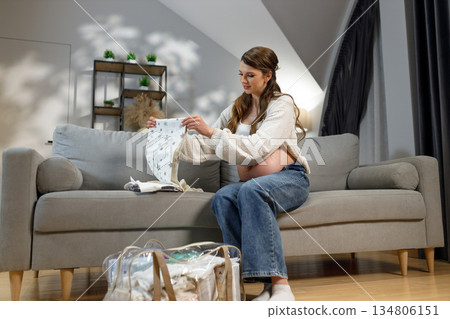 Expecting mother sits on sofa folding baby garments preparing for hospital arrival with bag nearby Expecting mother sits on sofa folding baby garments preparing for hospital arrival with bag nearby 134806151