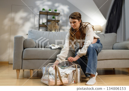 Young woman sitting on sofa, packing clothes into transparent duffel bag for travel or storage Young woman sitting on sofa, packing clothes into transparent duffel bag for travel or storage 134806153