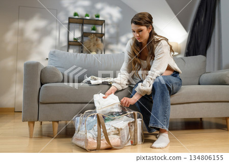 Young woman sits on sofa packing baby clothes into transparent bag, preparing for hospital visit or travel 134806155