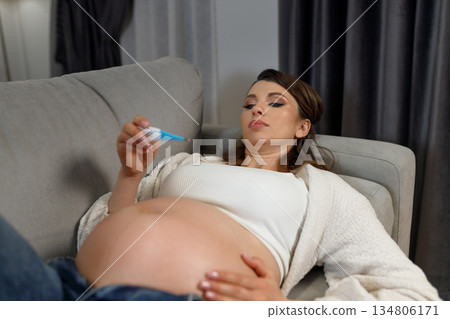 Pregnant woman lying on sofa holding thermometer checking her temperature feeling unwell during pregnancy 134806171
