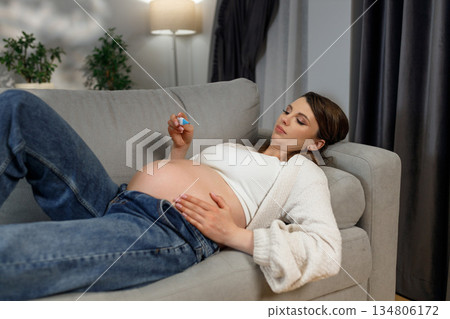 Pregnant woman resting on couch, holding small object, showing belly, wearing casual clothes, indoor setting with plants and curtains 134806172