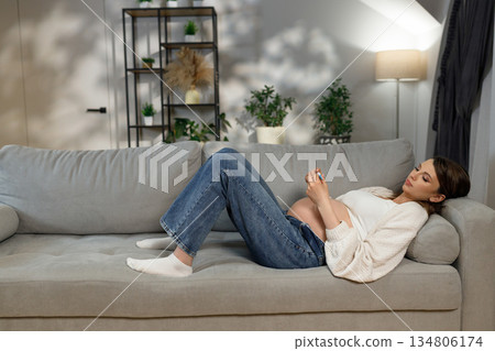 Pregnant woman reclines on a comfortable sofa, holding a mobile phone in her hands, with a serene expression 134806174