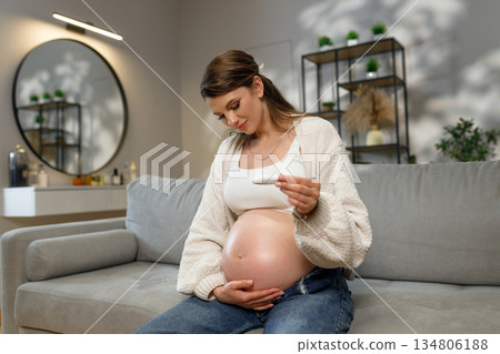 Pregnant woman with large belly sits on sofa holding pregnancy test, looking at it with anticipation and joy 134806188