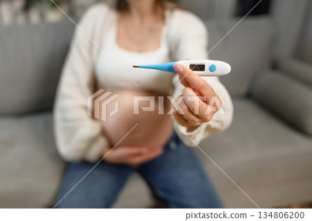 Pregnant woman sits on sofa holding digital thermometer showing normal temperature 36.5 degrees Celsius Pregnant woman sits on sofa holding digital thermometer showing normal temperature 36.5 degrees Celsius 134806200