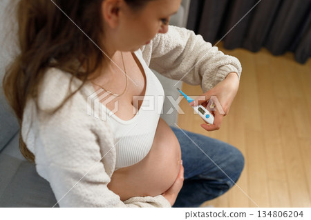 Pregnant woman holding digital thermometer checking her body temperature while sitting on couch, concerned about health during pregnancy Pregnant woman holding digital thermometer checking her body temperature while sitting on couch, concerned about health during pregnancy 134806204