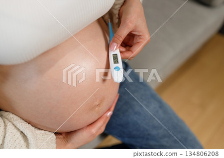 Woman holding digital thermometer near pregnant belly showing normal temperature reading for health monitoring 134806208