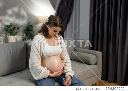 Pregnant woman sitting on sofa holding thermometer and touching belly, feeling unwell at home 134806211