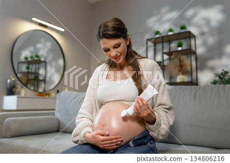 Expecting mother applies moisturizing cream to her belly while sitting on sofa, promoting skin elasticity and comfort during pregnancy Expecting mother applies moisturizing cream to her belly while sitting on sofa, promoting skin elasticity and comfort during pregnancy 134806216