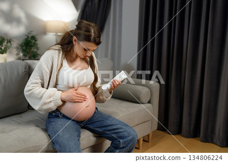 Pregnant woman sits on sofa applying cream to her belly for skin care during pregnancy 134806224