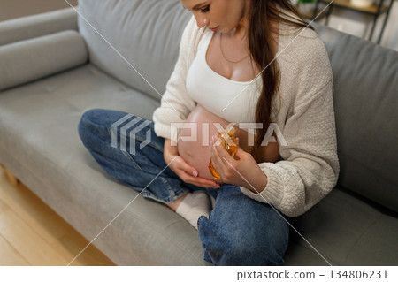 Expecting mother sits on sofa applying moisturizing oil to her pregnant belly for skin care during pregnancy Expecting mother sits on sofa applying moisturizing oil to her pregnant belly for skin care during pregnancy 134806231