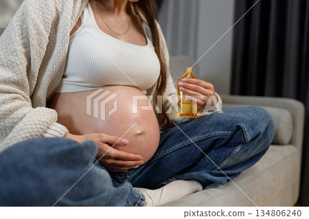 Pregnant woman holding bottle of oil for belly care while sitting on couch, expecting mother nurturing skin Pregnant woman holding bottle of oil for belly care while sitting on couch, expecting mother nurturing skin 134806240