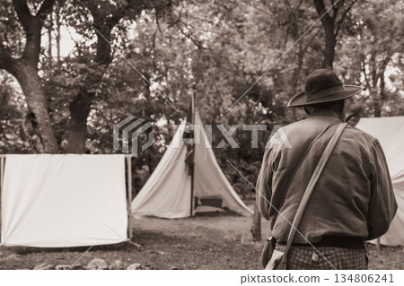 Southern reenactor in period attire stands before a Confederate flag and canvas tents at a historical event in the woods Southern reenactor in period attire stands before a Confederate flag and canvas tents at a historical event in the woods 134806241