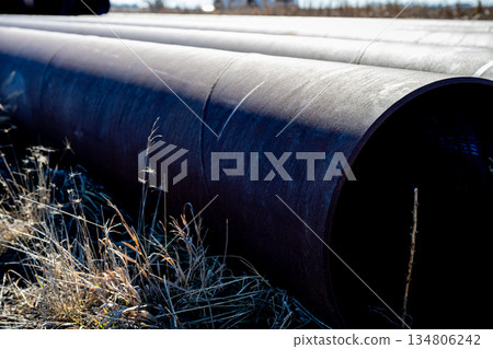 Large black pipes lie in a field near industrial buildings during a clear day 134806242