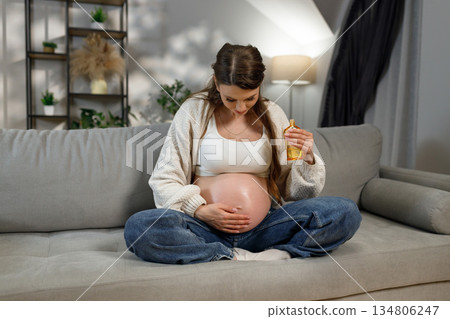 Pregnant woman sitting on sofa applying oil to her belly for stretch mark prevention and skin hydration during pregnancy 134806247