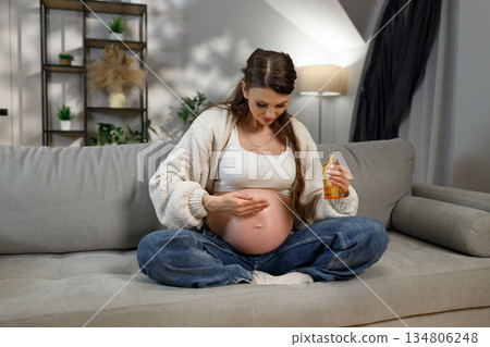 Expecting mother sits cross-legged on sofa applying moisturizing oil to her pregnant belly for skin care Expecting mother sits cross-legged on sofa applying moisturizing oil to her pregnant belly for skin care 134806248