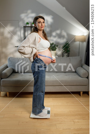 Pregnant woman stands on scale measuring her belly circumference with tape measure at home 134806253