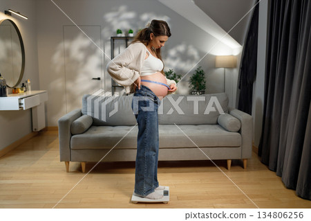 Pregnant woman stands on scale measuring her belly circumference with tape measure at home 134806256